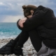 A woman practicing Somatic Experiencing exercises on a rocky beach, sitting with her knees drawn to her chest and her arms wrapped around them.