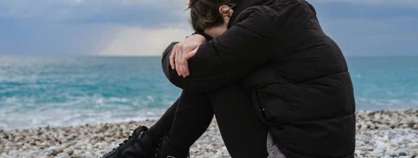 A woman practicing Somatic Experiencing exercises on a rocky beach, sitting with her knees drawn to her chest and her arms wrapped around them.