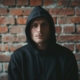 A man wearing a dark hoodie stands against a textured brick wall, staring intensely directly at the camera with a serious expression showing the concealment associated with pathological lying.