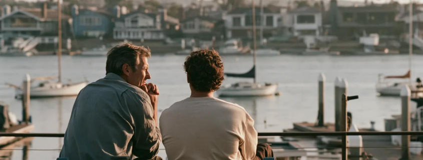 Two men having a supportive conversation on a bench overlooking a harbor.